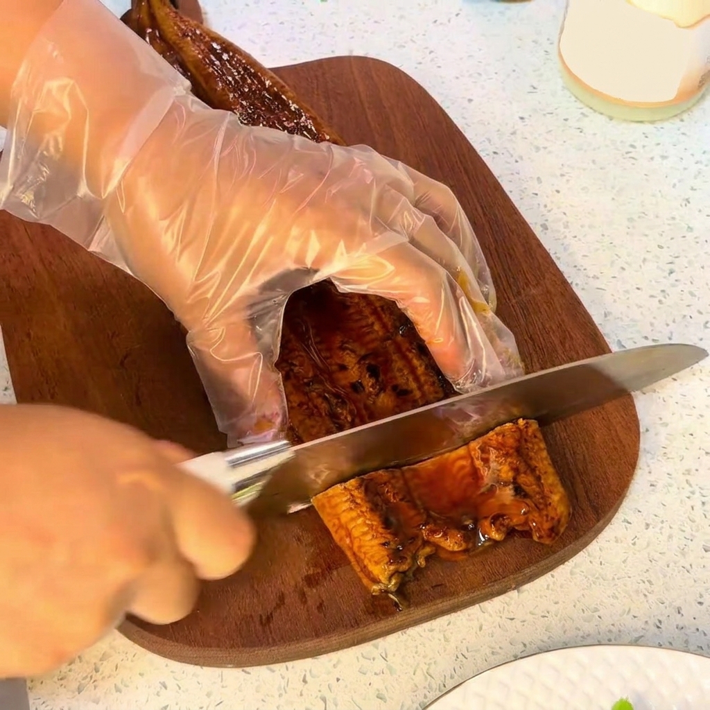 A knife cutting through a piece of grilled eel on a wooden cutting board.