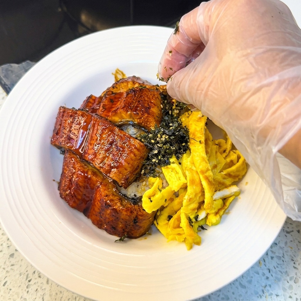 A hand wearing a plastic glove sprinkling shredded nori and sesame seeds over a bowl of grilled eel and rice with egg strips.