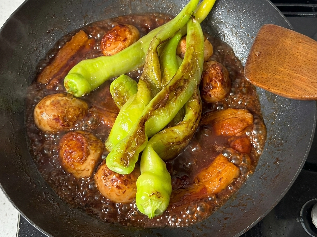 Green peppers simmering in the dark, rich sauce with pork belly and hard-boiled eggs in a pan.