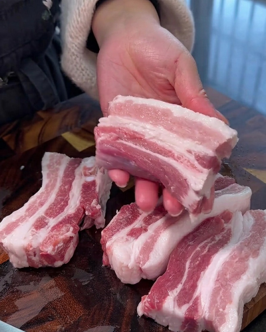 Raw pork belly being sliced into thick, uniform strips on a wooden cutting board.