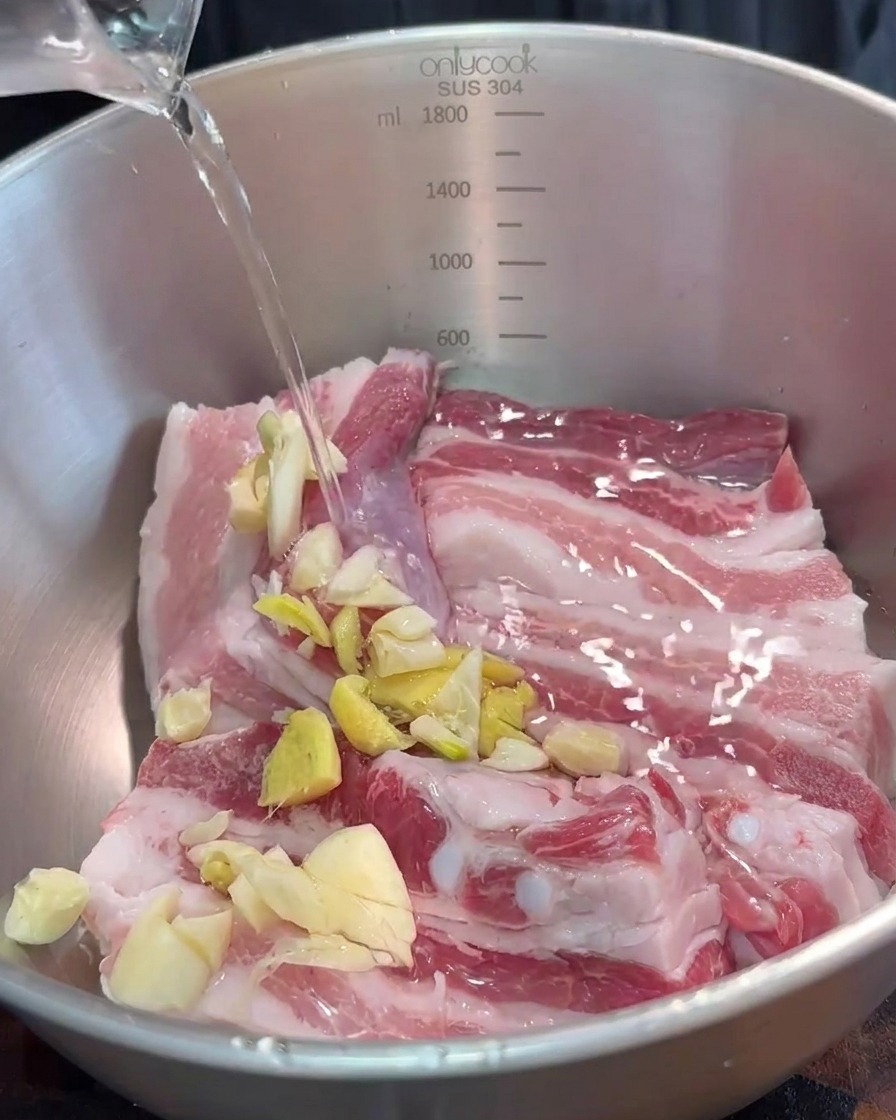 A bowl containing pork belly strips being seasoned with minced aromatics and red fermented bean curd sauce.