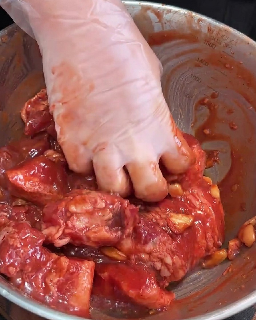 Gloved hands massaging the marinade into the raw pork belly in a stainless steel mixing bowl.