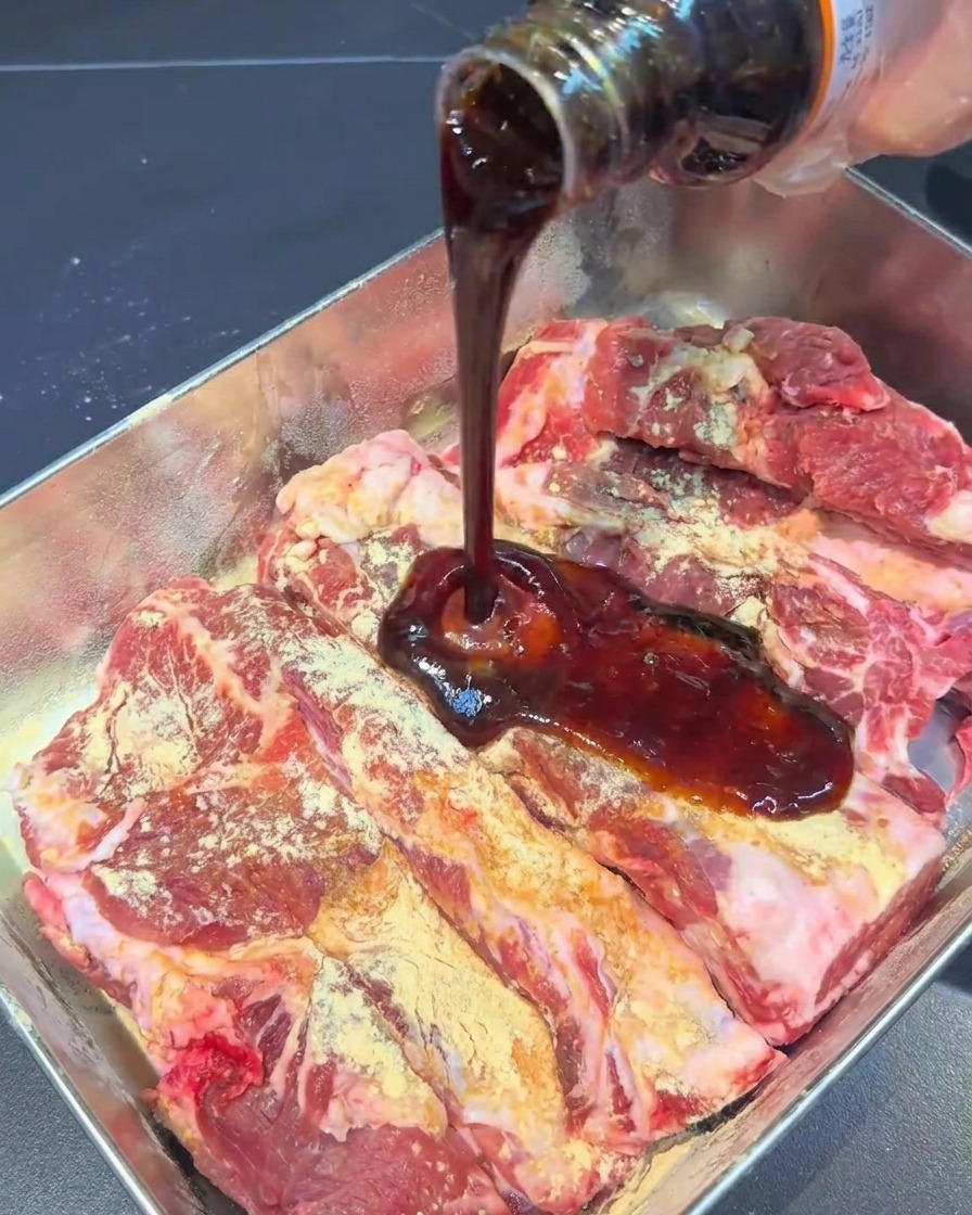 Dark marinade sauce being poured from a glass bottle onto seasoned pork ribs in a metal tray.
