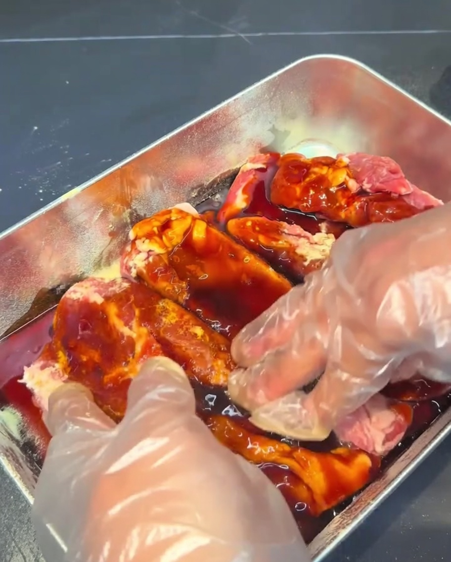 Pork ribs being massaged and coated in a dark marinade sauce inside a metal container.