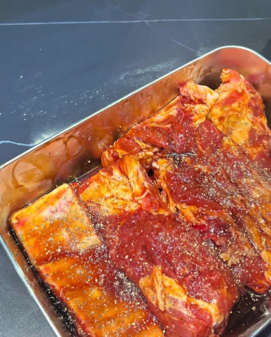 Close up of raw pork ribs completely coated in a dark marinade and sprinkled with coarse black pepper flakes.