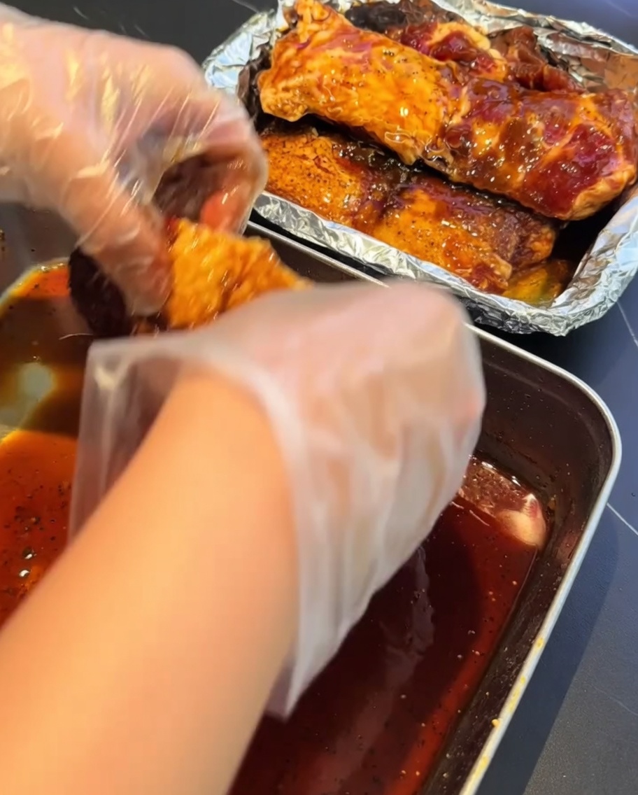 Gloved hands transferring marinated pork ribs into an aluminum foil-lined container for steaming.