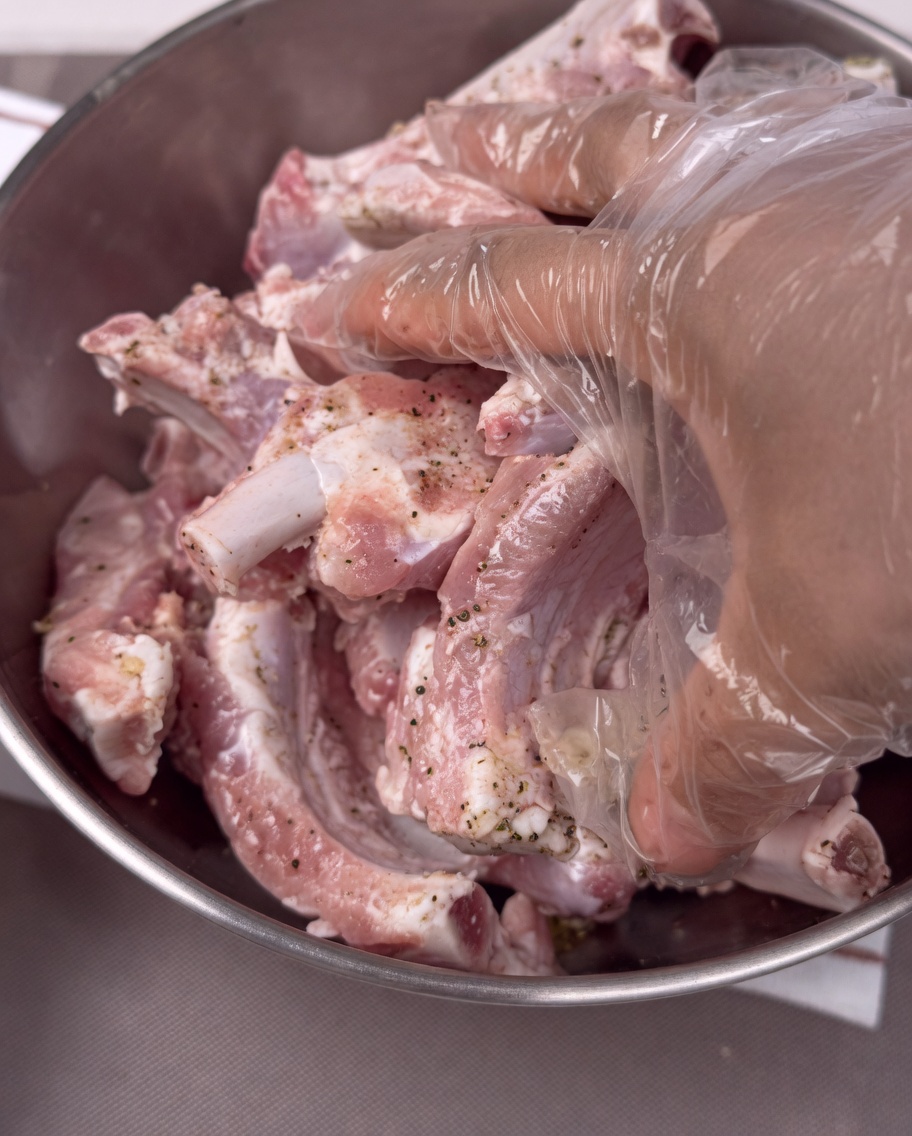Hands wearing plastic gloves mixing seasoned pork ribs in a stainless steel bowl.