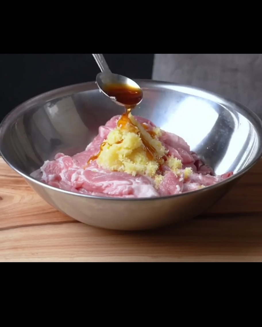 A spoon pouring dark syrup over raw pork ribs topped with minced aromatics in a metal bowl.