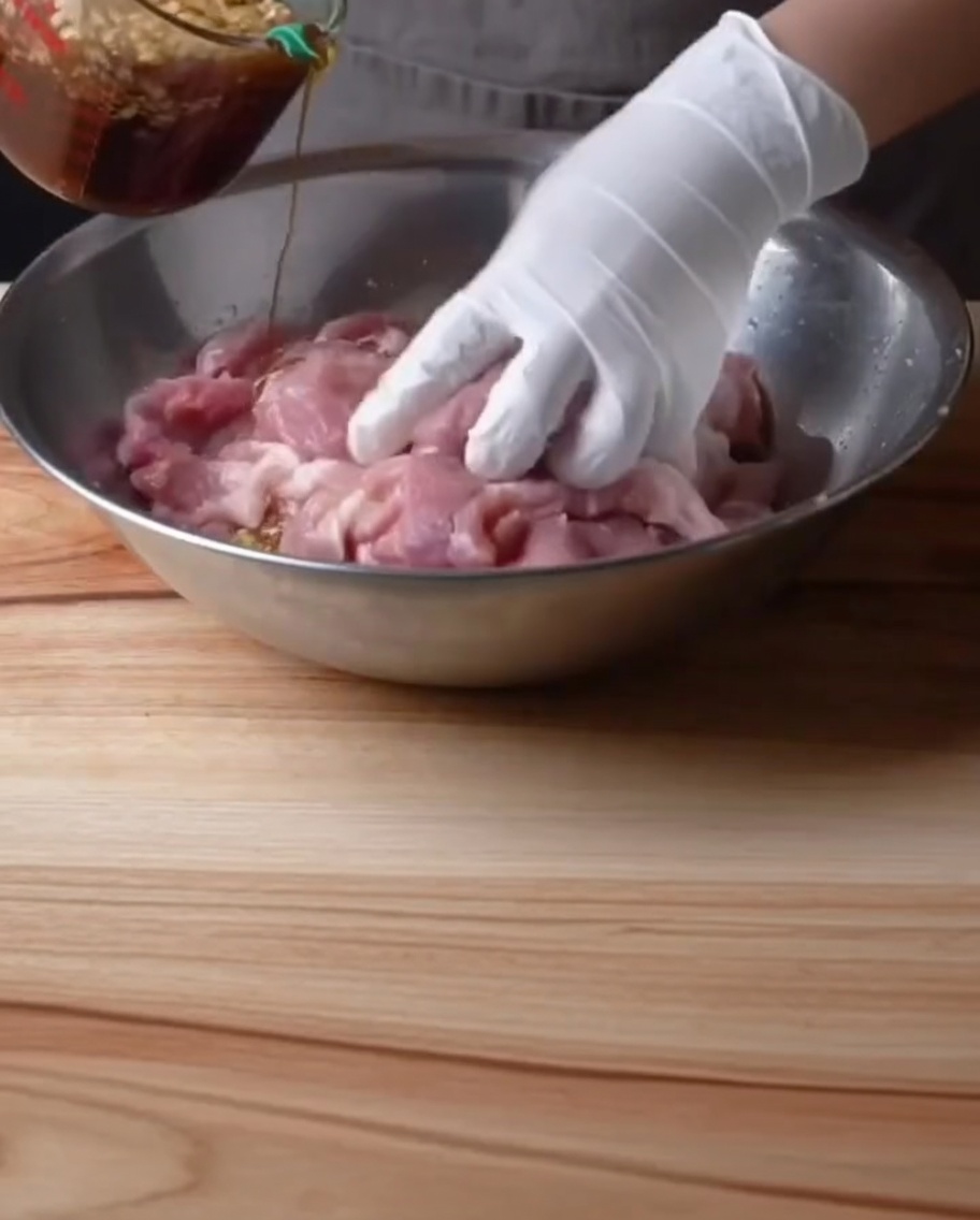 A gloved hand pouring dark, seasoned marinade from a measuring cup over raw pork ribs in a large metal bowl.