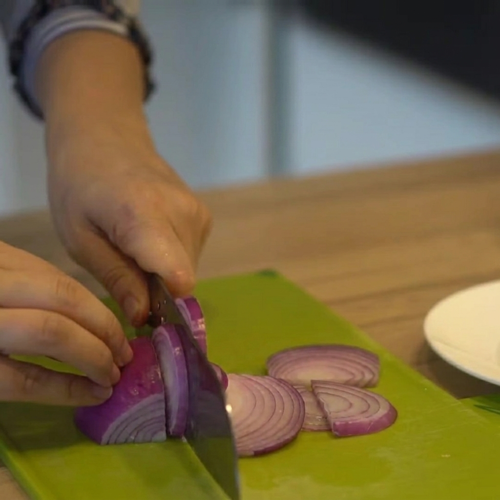 Red onion being cut into thin slices on a green cutting board.