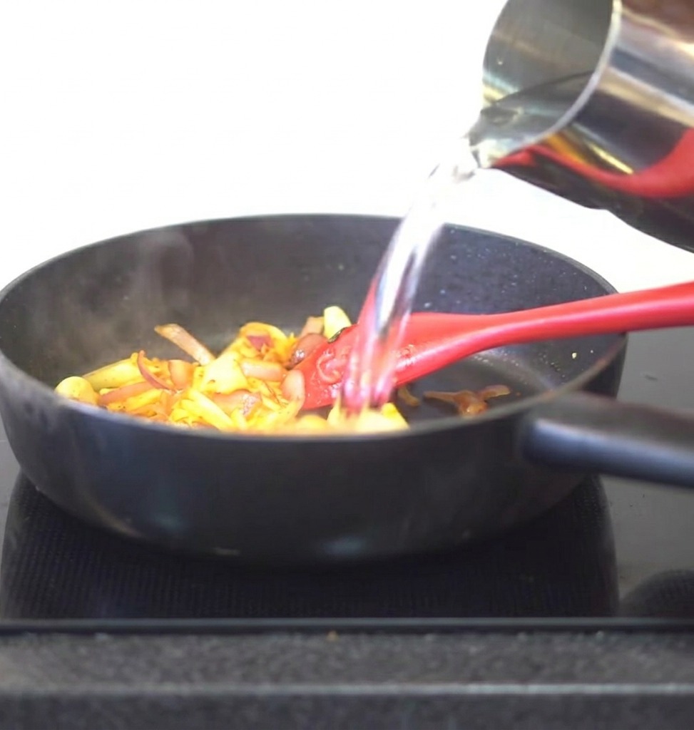 Water pouring from a metal pitcher into a pan containing stir-fried kimchi and aromatics.