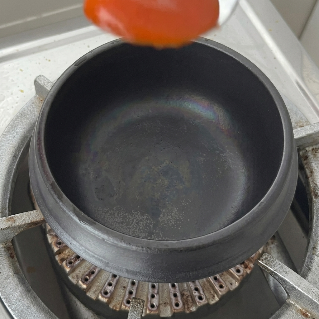 A spoonful of spicy red chili sauce being placed into the bottom of a dry black clay pot on a stovetop.