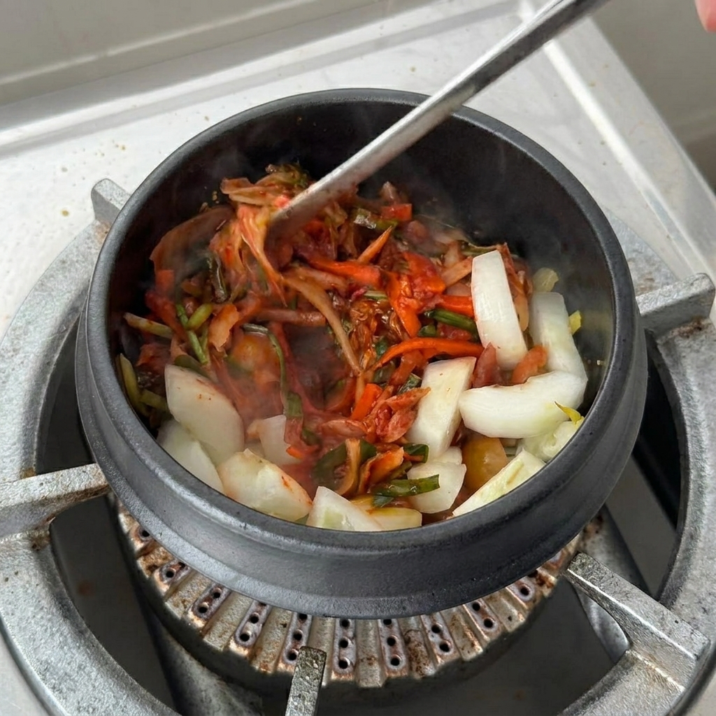 A large cup of red kimchi soup base being poured into the clay pot over the sautéed kimchi and onions.