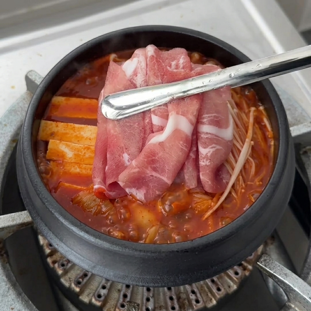 Fresh slices of raw pork being arranged on top of the simmering Kimchi Jjigae ingredients.
