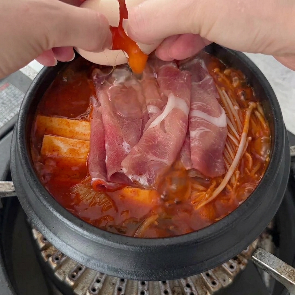 Two fresh eggs being cracked onto the bubbling surface of the Kimchi Jjigae, with the broth and ingredients visible below.