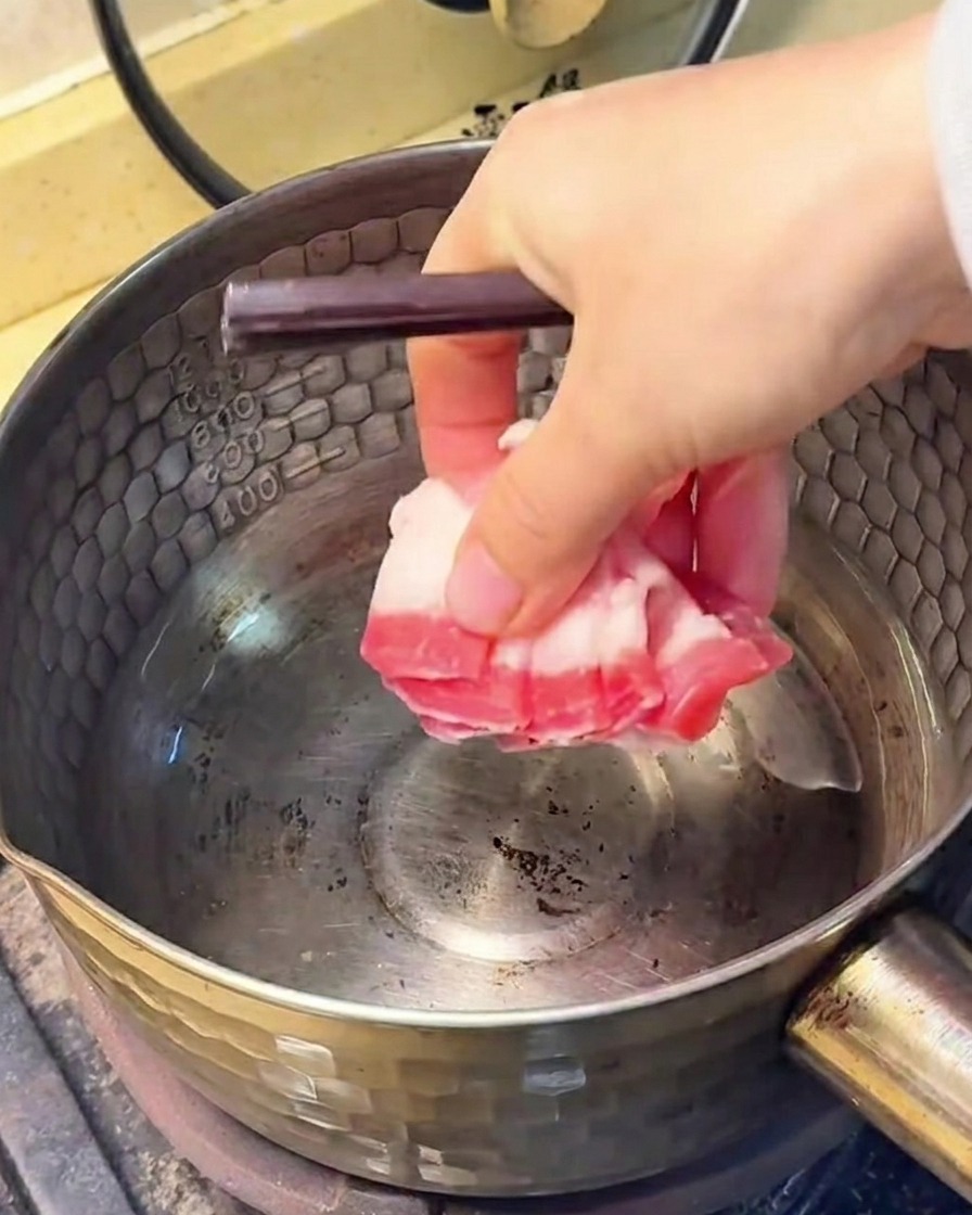 Slices of pork belly frying in a stainless steel pot, with rendered fat visible at the bottom.