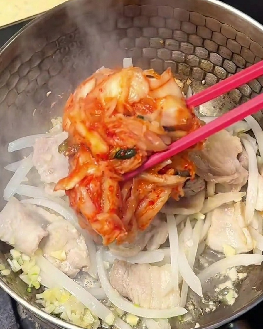 Red, seasoned kimchi being stirred into the pork and onion mixture in the pot.