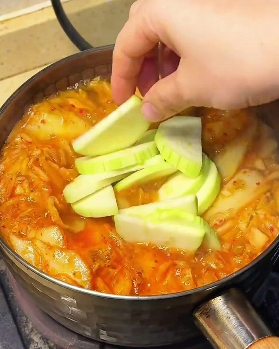 Vegetables, including sliced chayote, bean sprouts, and enoki mushrooms, being added to the simmering soup.