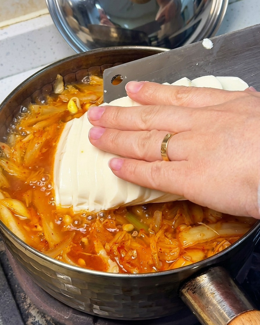 A close-up of a hand gently placing sliced silken tofu into the bubbling red kimchi soup.