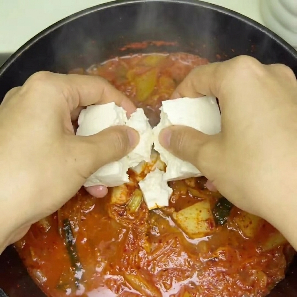 Fresh tofu cubes being placed into the bubbling red kimchi soup.