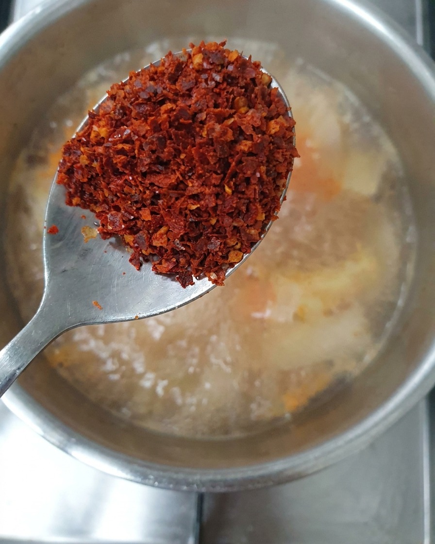 A spoonful containing a mix of coarse and fine red chili flakes held over the stew.