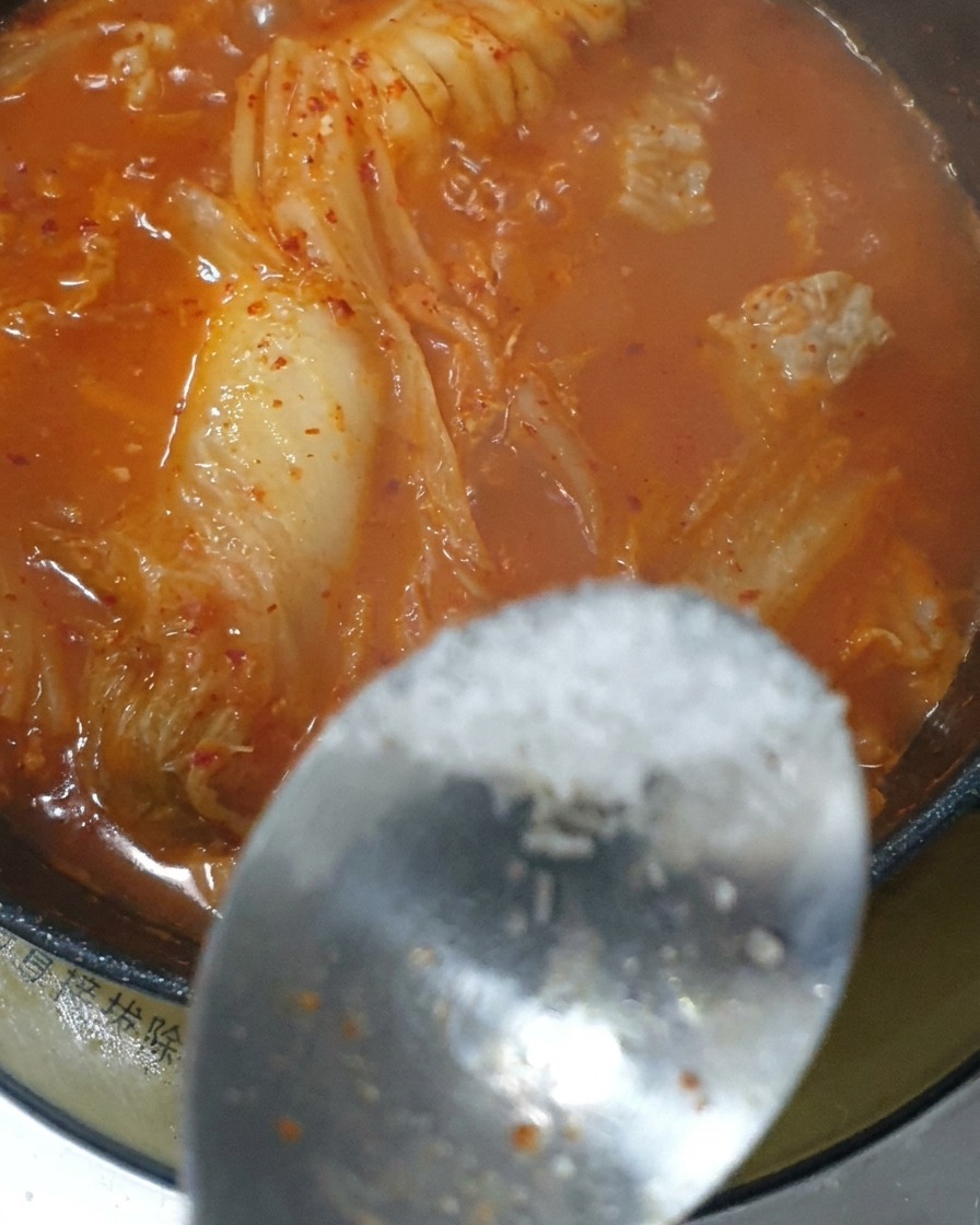 A spoonful of seasoning being added to the simmering red kimchi stew.