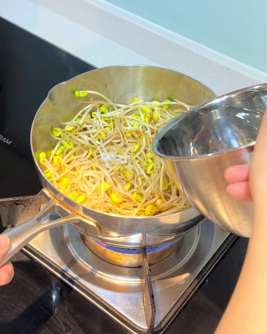 Water being poured from a stainless steel bowl into a pot containing bean sprouts and other stir-fried ingredients.