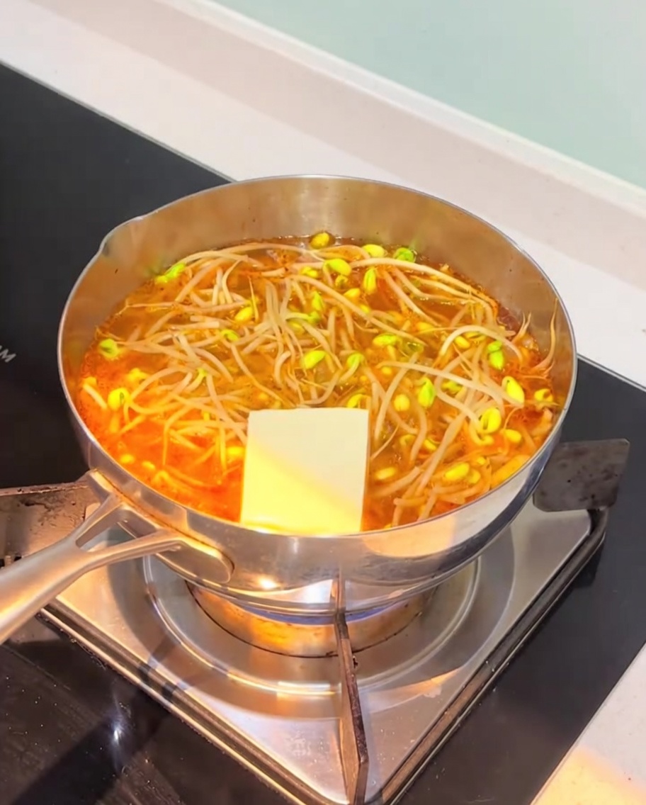 Slices of white tofu and fish cakes being added to the simmering orange kimchi soup base.