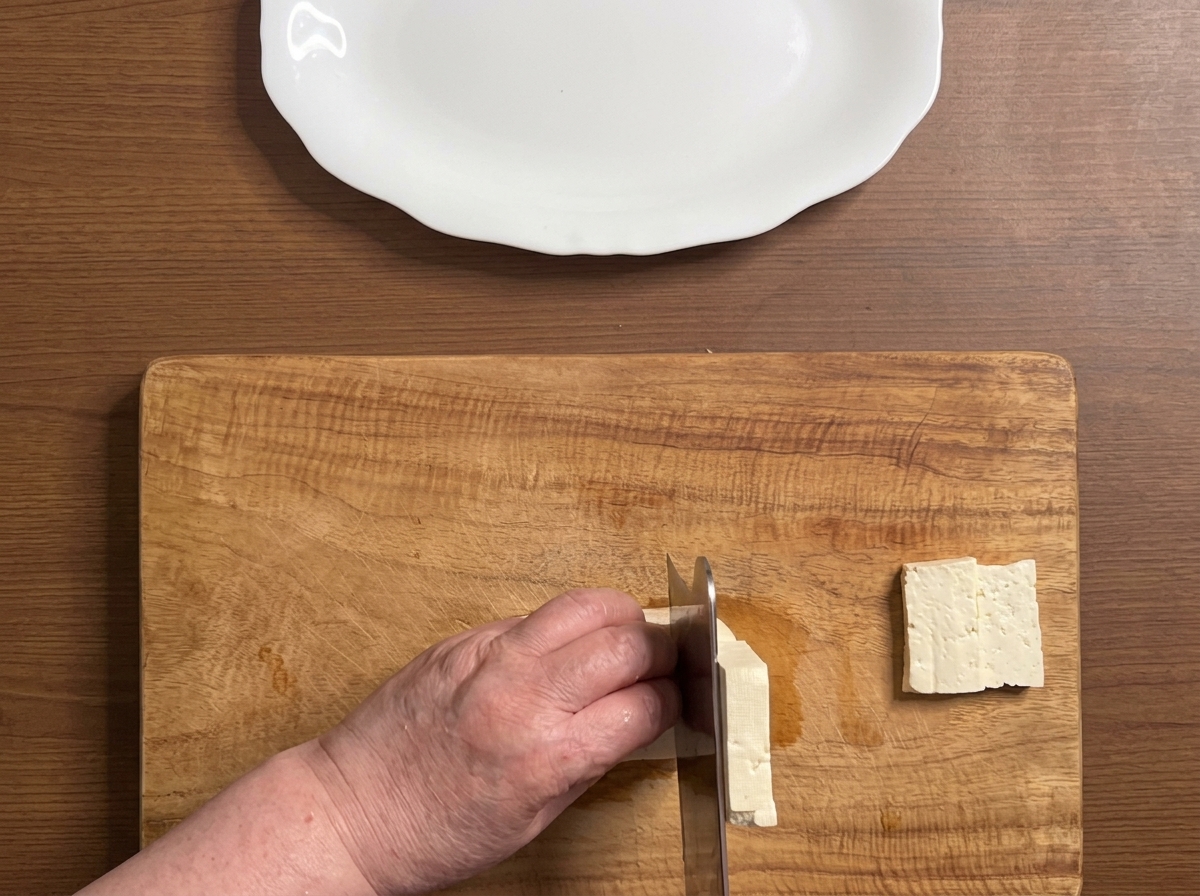 Tofu block being cut into thick, neat slices on a wooden cutting board.
