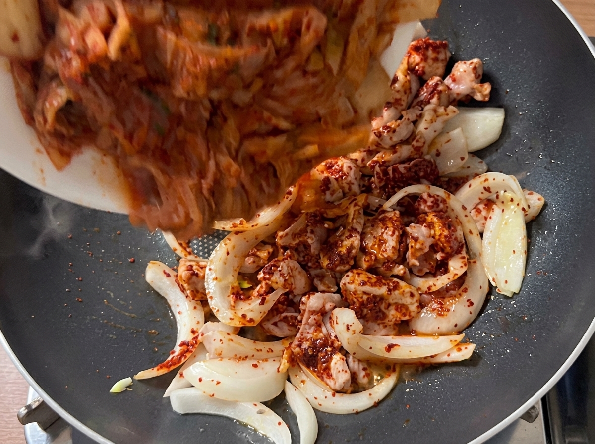 Bite-sized kimchi pieces being poured into the pan with the browned pork and onion mixture.