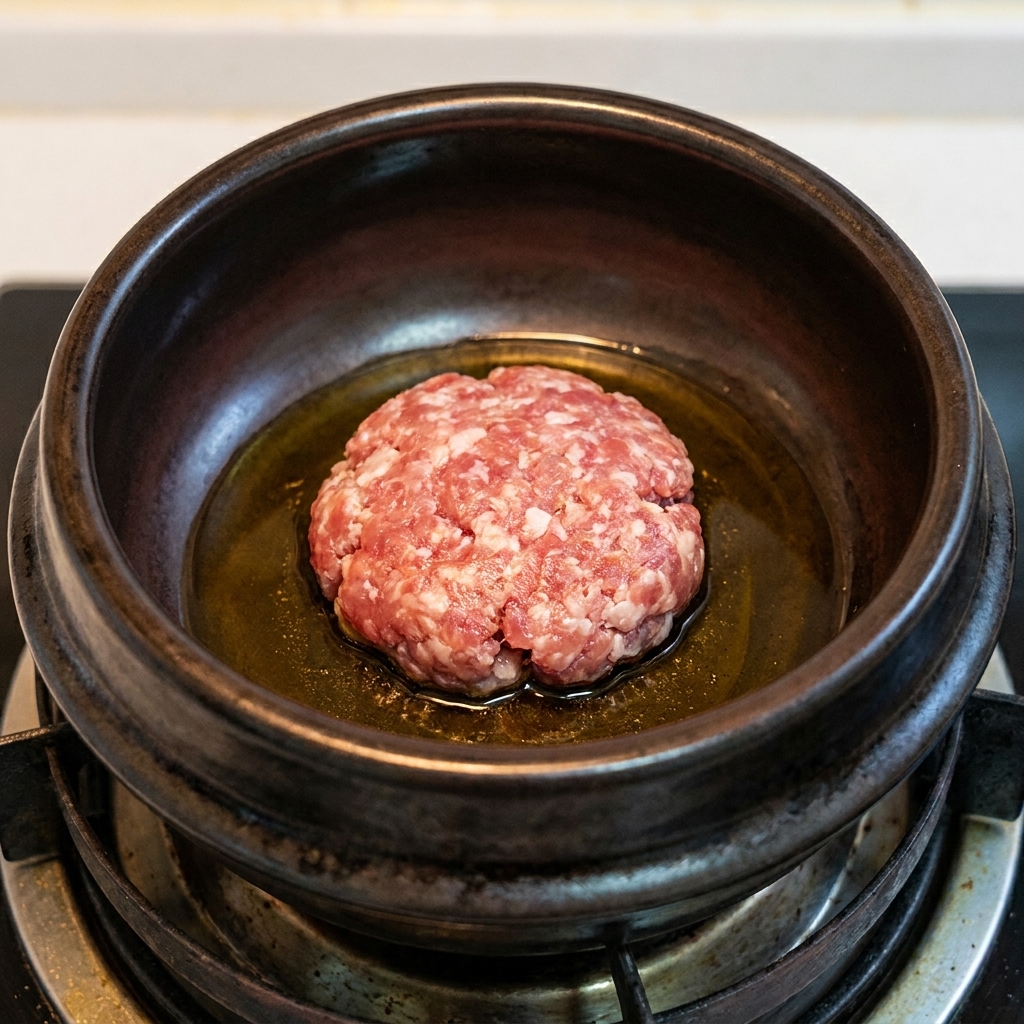A ball of ground pork browning in hot sesame oil inside a dark earthenware pot.