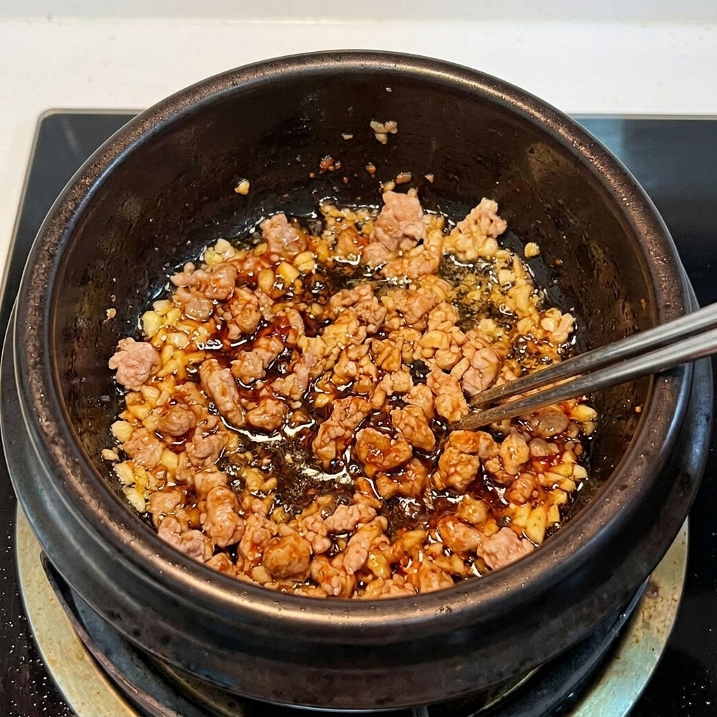 Sliced kimchi and ground pork being stirred together in an earthenware pot, sautéing until fragrant.