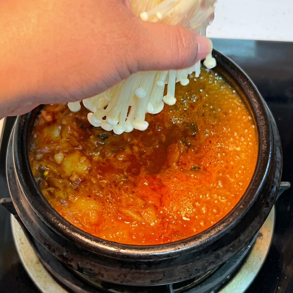 A hand placing a bundle of fresh enoki mushrooms into the bubbling, red kimchi stew.