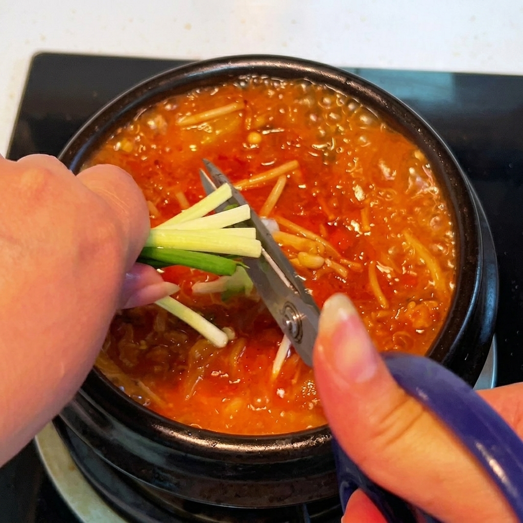 Fresh green onions being cut directly over the simmering pot of kimchi stew using a pair of kitchen scissors.