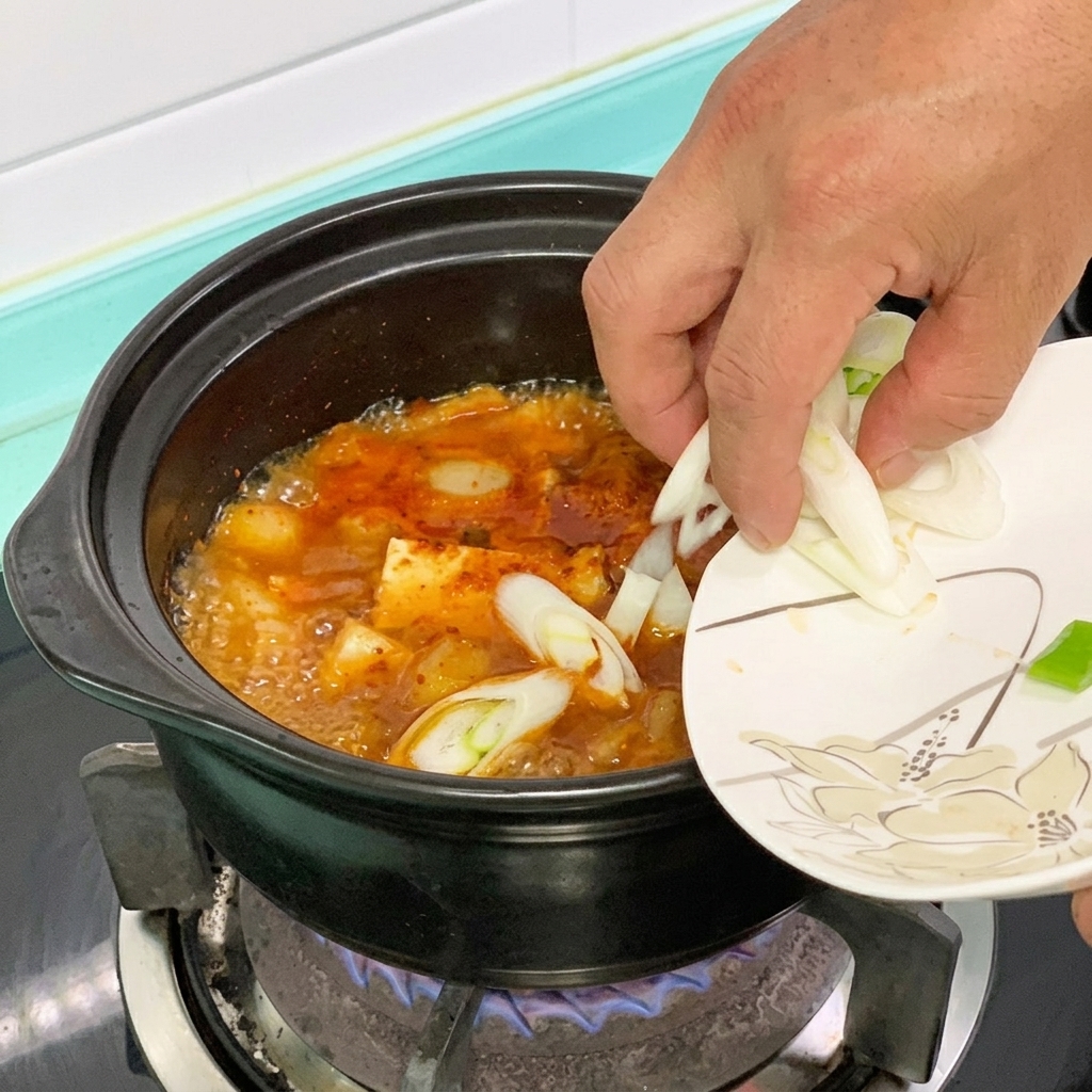 Green chili and green onion being dropped into a pot of red-hued tofu stew.
