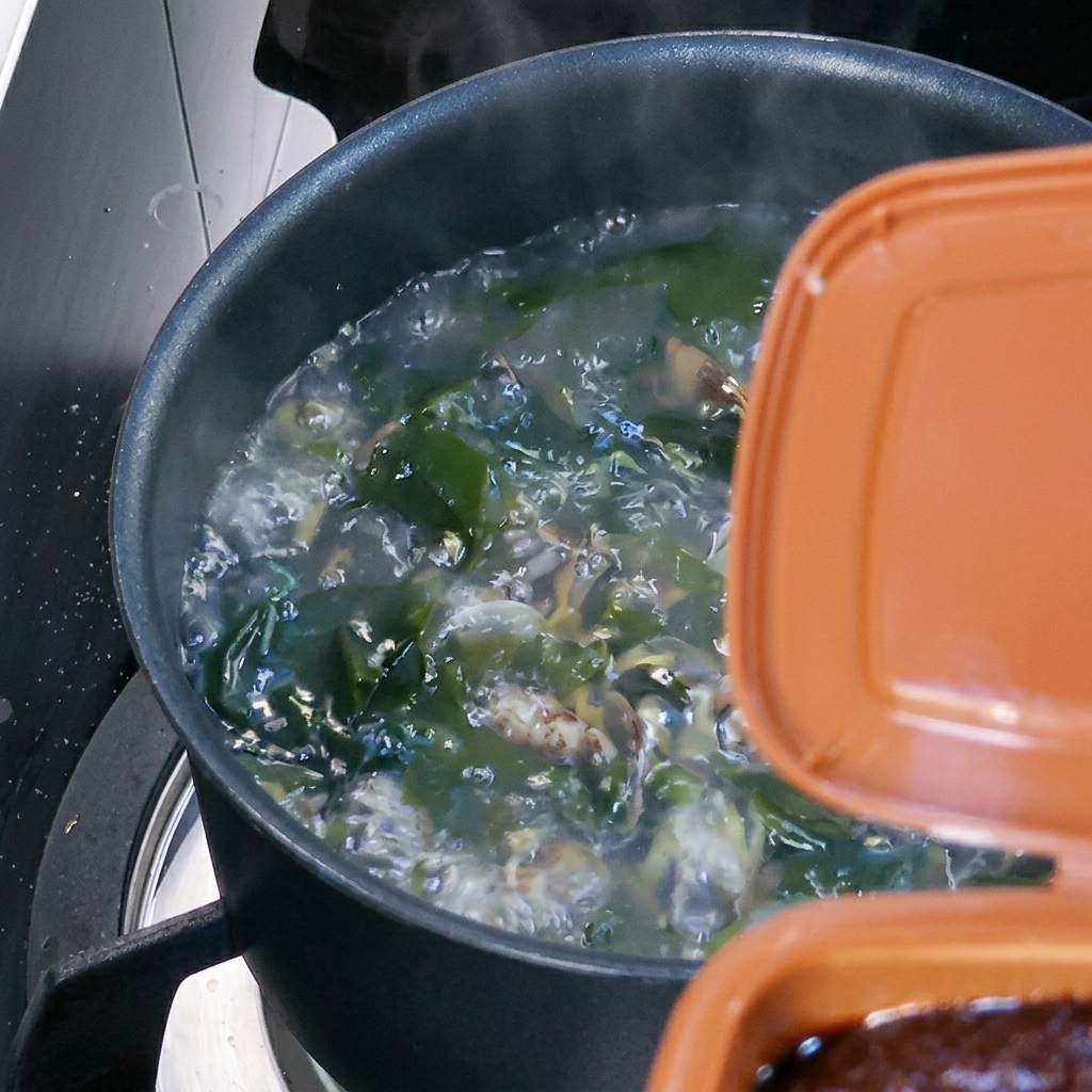 A spoonful of brown Korean soybean paste being stirred into the boiling soup.
