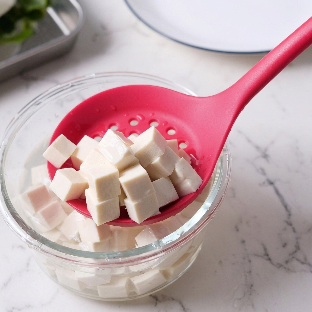 Cubed tofu being lifted out of a bowl of water using a pink slotted spoon.