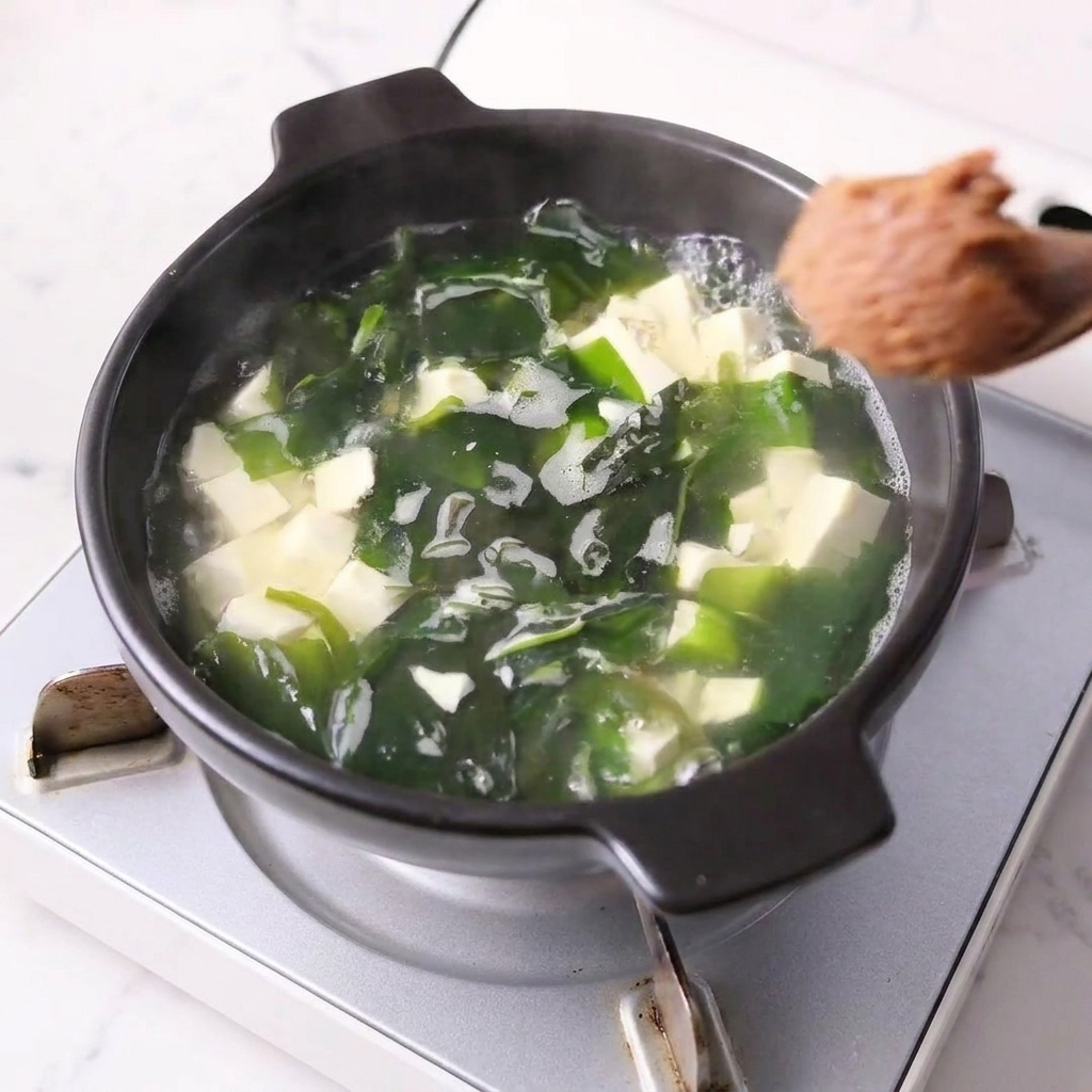 A spoonful of miso paste being stirred into a pot with tofu and wakame.