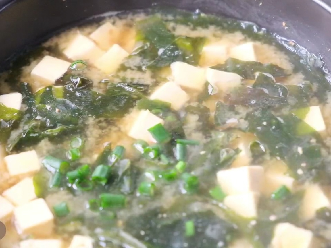 A close-up of a pot of hot miso soup with cubes of tofu, seaweed, and fresh green onions sprinkled on top.