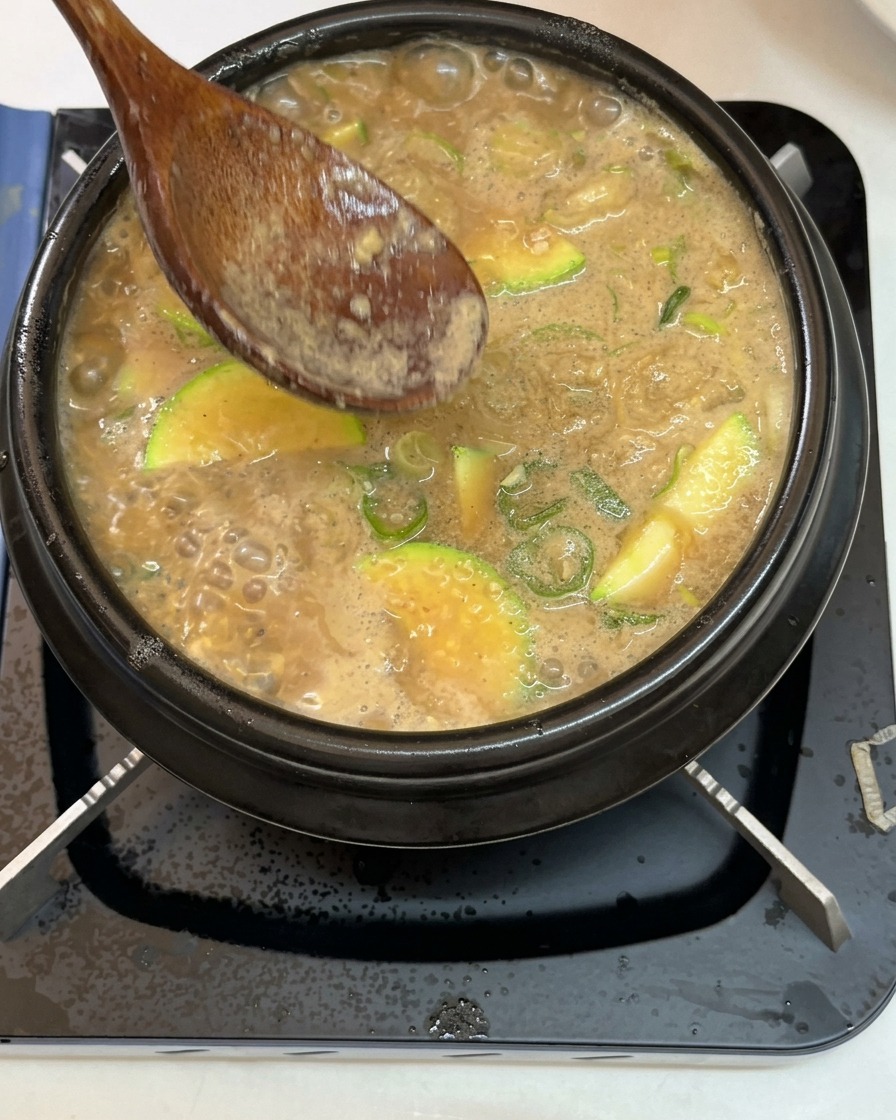 A wooden spoon removing foam from the surface of the bubbling soybean paste stew.