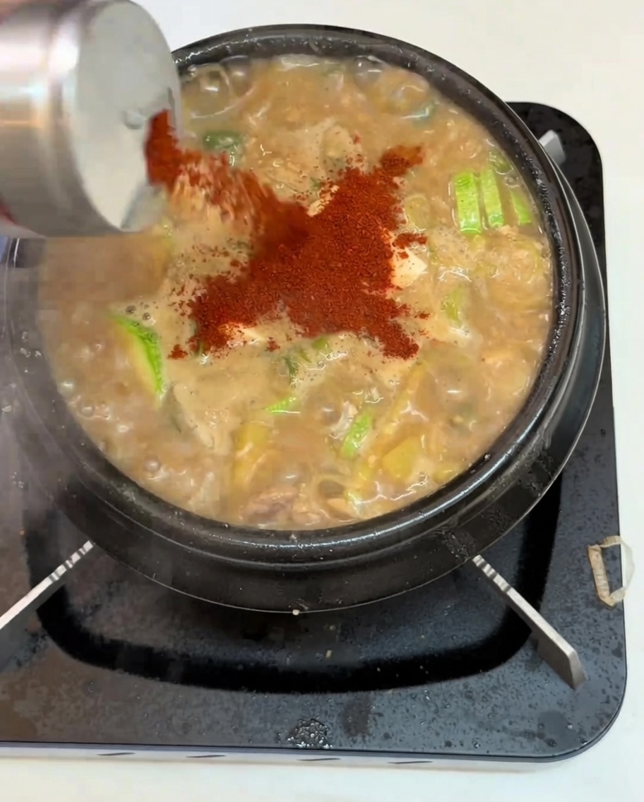 A close-up of red chili powder being shaken from a container onto a pot of bubbling, savory soybean paste stew containing tofu, zucchini, and vegetables.
