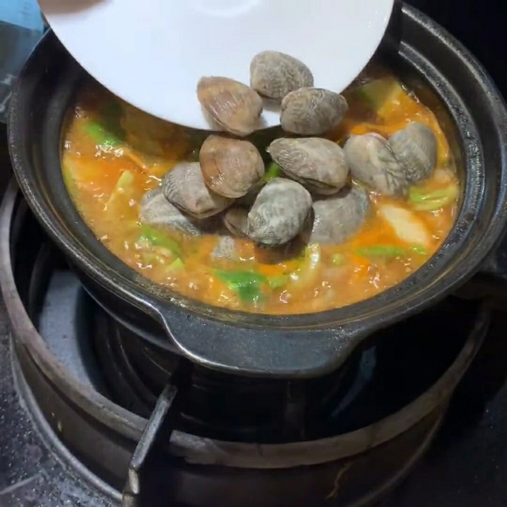 Fresh clams and soft tofu cubes being carefully placed into the bubbling Korean soybean paste stew.