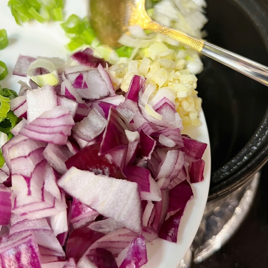 Fresh green onions, garlic, and red onion being mixed into the sautéed pork belly in the pot.