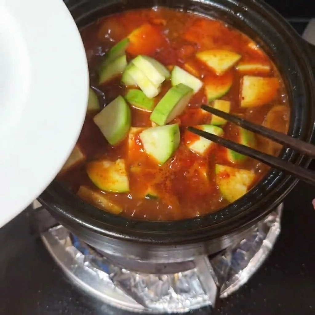 Slices of zucchini and cubes of soft silken tofu being placed into a bubbling red soup.