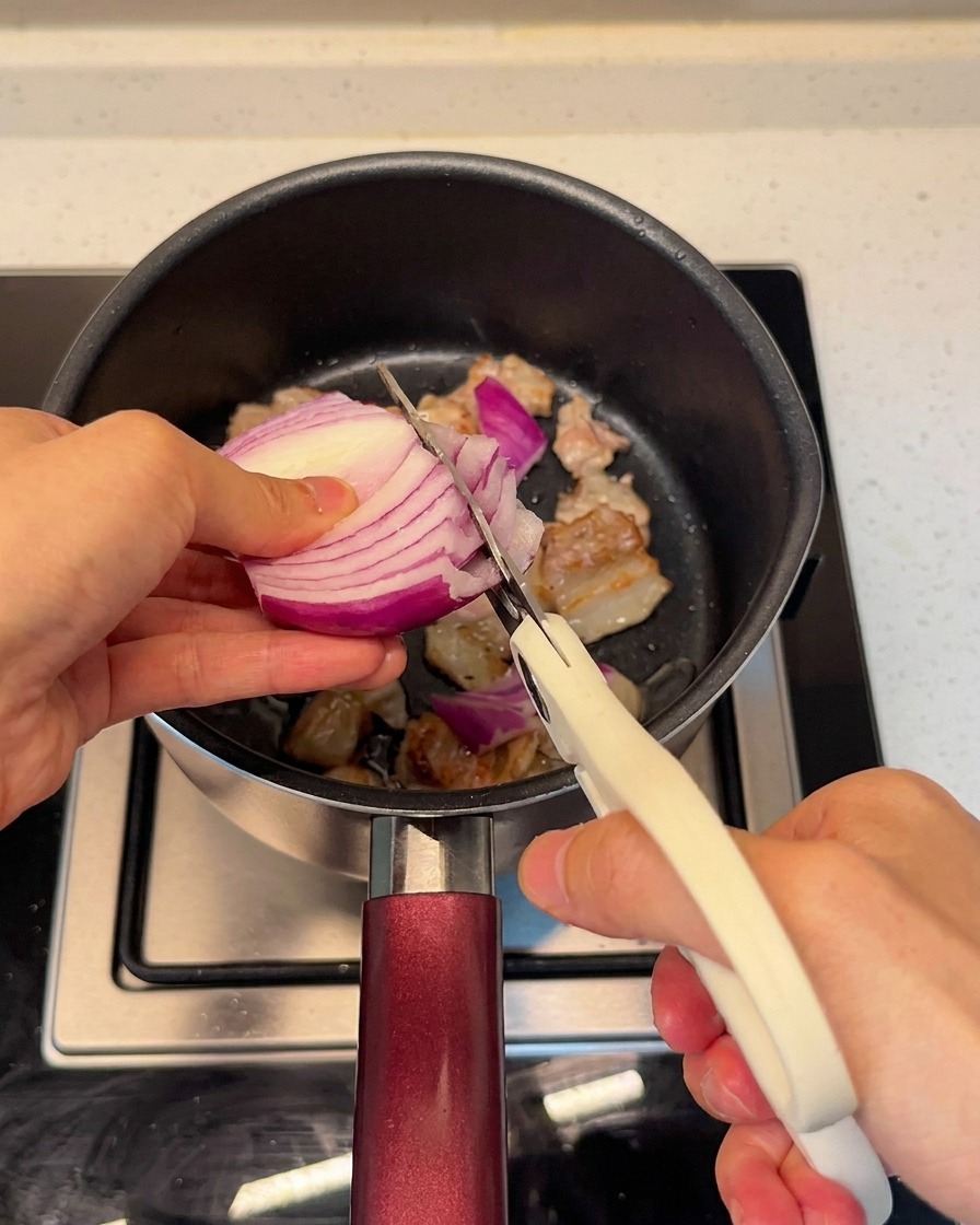 Slicing fresh red onion directly into the pot with the pork belly using kitchen scissors.