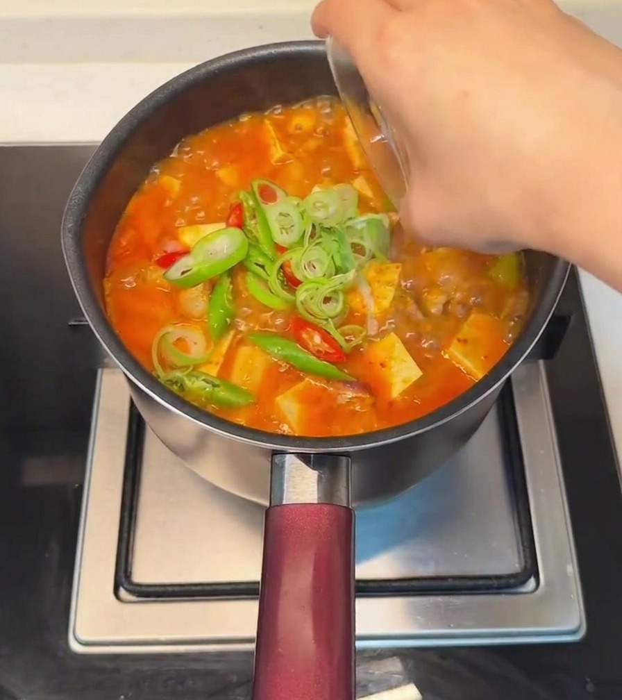 A hand scattering sliced green onions and fresh red and green chili peppers over the finished tofu stew.