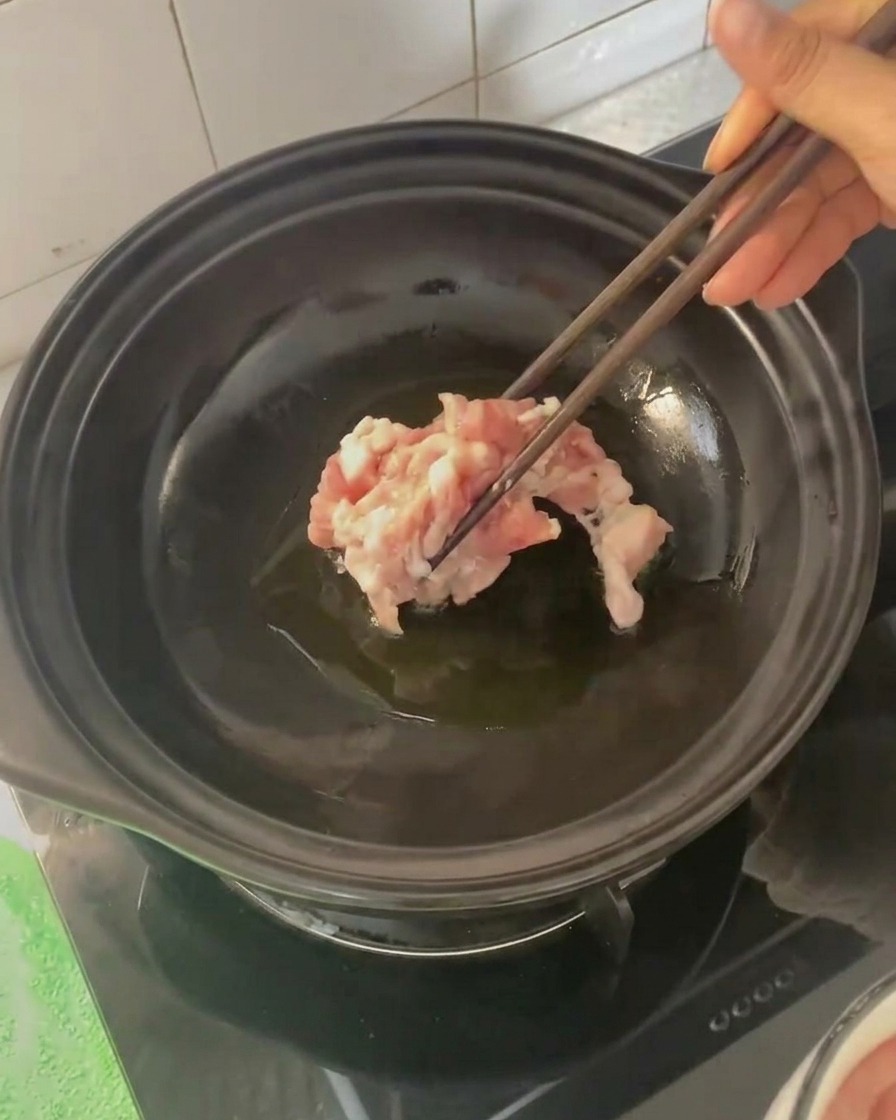 Pork belly slices being added to the hot clay pot with chopsticks.