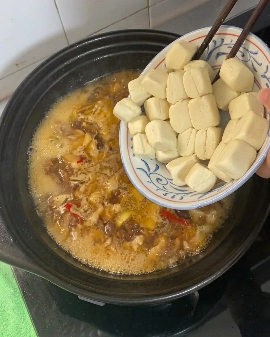 Small tofu cubes being added to a bubbling, golden-brown braising liquid in a clay pot.