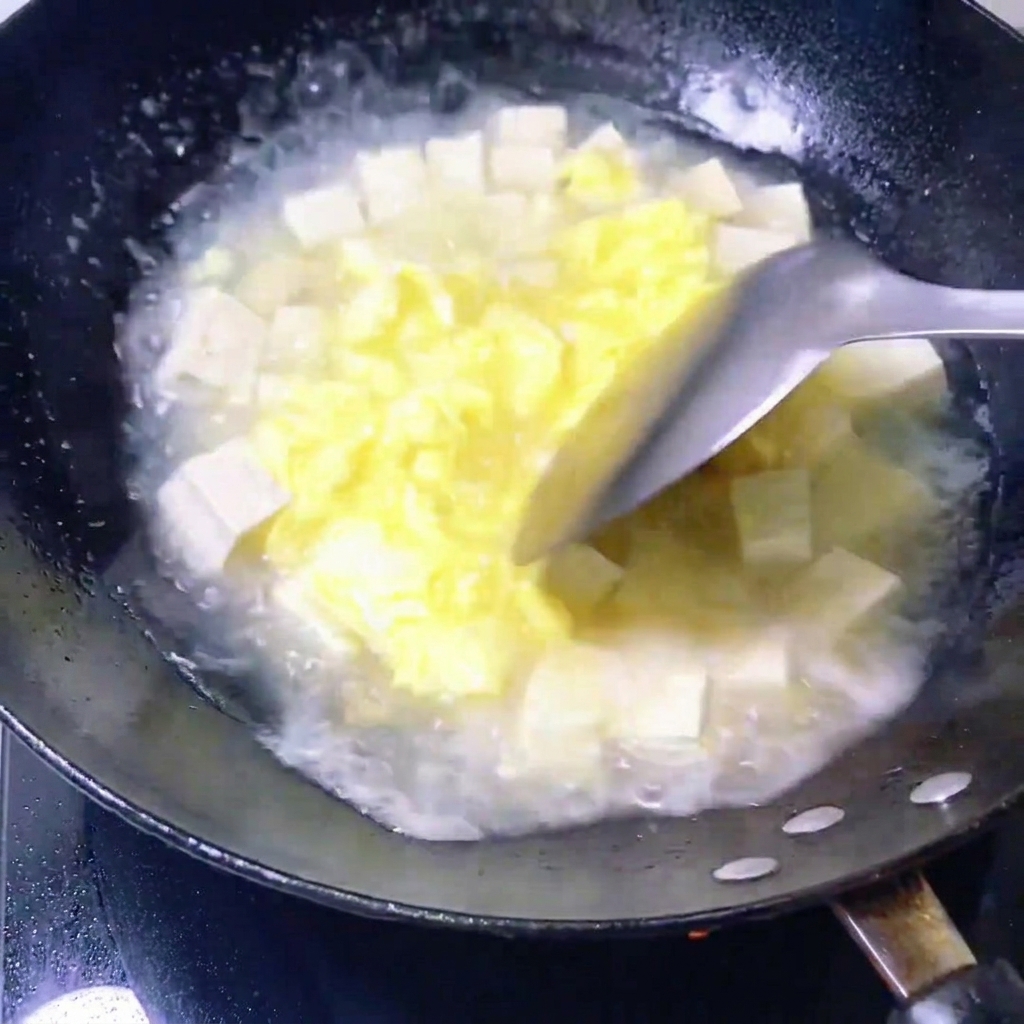 Fresh clams being placed into a pan containing a simmering mixture of egg broth and tofu cubes.