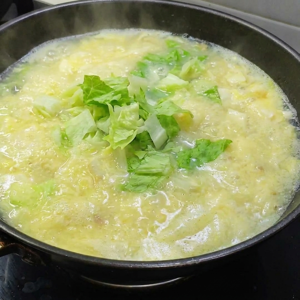 Fresh green lettuce leaves being added to a pot of simmering golden salted egg soup.